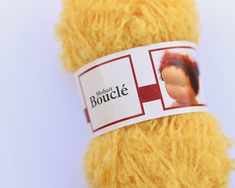 K&ouml;nnte beinhalten: Ein Kn&auml;uel aus leuchtend gelbem Mohair-Boucl&eacute;-Garn mit strukturierter, lockiger Optik. Ein wei&szlig;es Papieretikett umschlie&szlig;t die Mitte und zeigt den Text Mohair Boucl&eacute; sowie eine kleine Abbildung eines Puppenkopfes mit rotbraunem Haar.