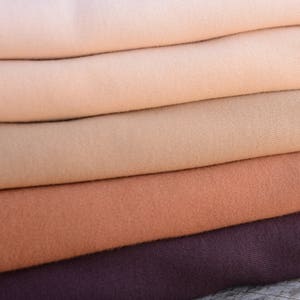 Op de afbeelding: Een stapel gevouwen stof in een reeks neutrale kleuren, van lichtbeige tot diepbruin. De stof lijkt zacht en mogelijk gebreid of geweven, met zichtbare textuur. De lagen zijn netjes opgestapeld en tonen de kleurvariaties.