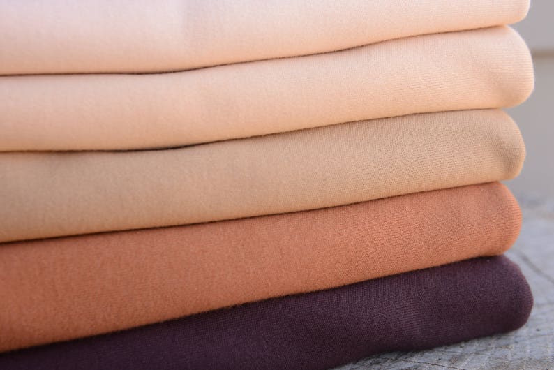 Op de afbeelding: Een stapel gevouwen stof in een reeks neutrale kleuren, van lichtbeige tot diepbruin. De stof lijkt zacht en mogelijk gebreid of geweven, met zichtbare textuur. De lagen zijn netjes opgestapeld en tonen de kleurvariaties.