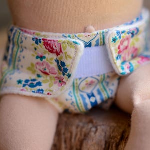 May include: Close-up of a cloth diaper with a floral pattern. The diaper features a cream-colored background with pink, red, and blue floral designs. A white Velcro closure is visible. The diaper is on a doll.