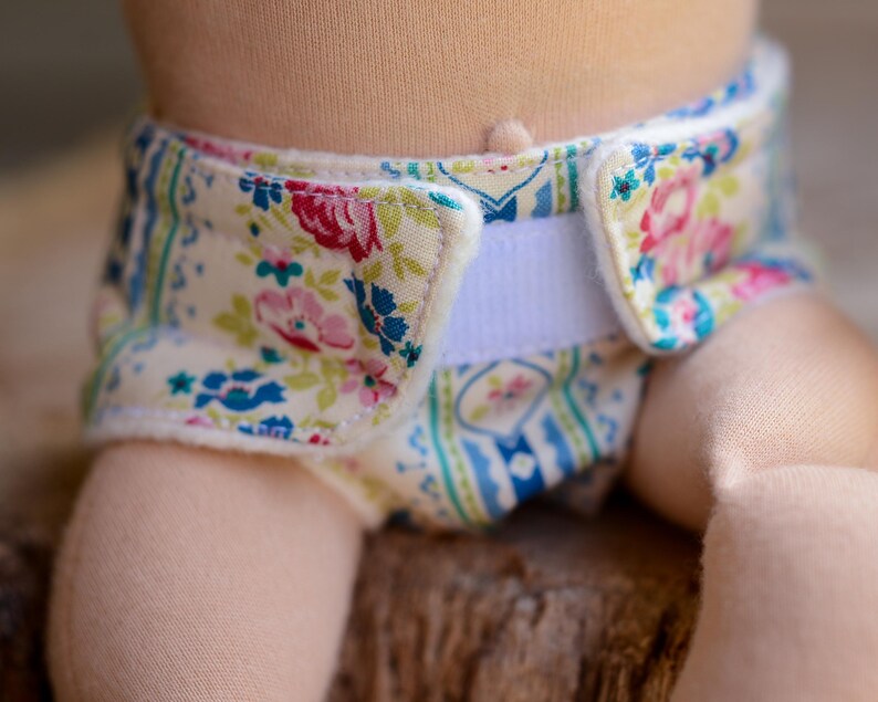May include: Close-up of a cloth diaper with a floral pattern. The diaper features a cream-colored background with pink, red, and blue floral designs. A white Velcro closure is visible. The diaper is on a doll.