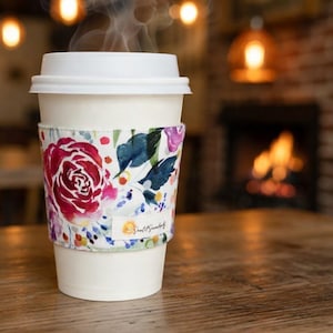 Puede incluir: Un vaso de café desechable blanco con tapa de plástico blanca y una funda de tela con un estampado floral de acuarela en tonos rojos, rosas, azules y verdes. Una pequeña etiqueta blanca con el texto Small & Sunny aparece en la funda.