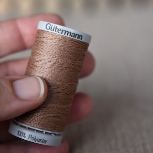 May include: A spool of brown thread with the brand name "Gütermann" and the text "100% Polyester" printed on the label.