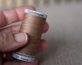 May include: A spool of brown thread with the brand name "Gütermann" and the text "100% Polyester" printed on the label.
