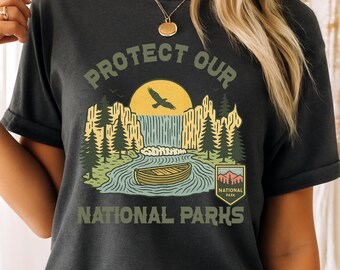 Puede incluir: Una camiseta gris oscuro con un diseño gráfico de estilo vintage que presenta una cascada, una canoa, un pájaro y árboles. El texto "Protect Our National Parks" está impreso en la camiseta.