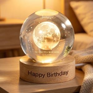 Puede incluir: Una esfera de vidrio transparente con una imagen detallada de la luna en su interior, sobre una base de madera. La base está grabada con "Happy Birthday". La esfera está iluminada desde dentro, y el texto "THE NIGHT YOU WERE BORN 05.12.2026" es visible.
