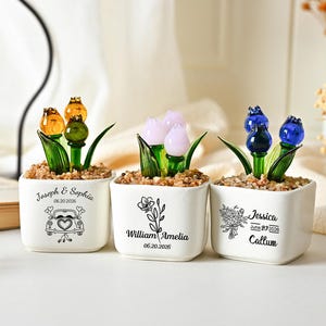 Peut inclure: Trois pots en céramique blanche carrés contenant des fleurs en verre orange, rose et bleu. Chaque pot présente des textes et graphiques imprimés en noir, incluant des noms comme Joseph & Sophia, William & Amelia et Jessica & Callum avec dates.