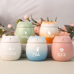 Puede incluir: Cinco tarros de cerámica redondeados con tapas en forma de gato en colores pastel: blanco, verde salvia, degradado naranja, degradado azul y rosa. Cada tarro tiene un gráfico blanco de una huella o silueta de gato y un nombre: Katrina, Emily, Gladys, Blake y Beata.