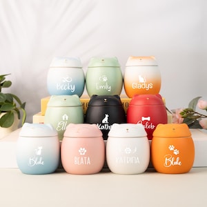 Può includere: Una collezione di urne a forma di gatto in vari colori, tra cui blu, verde, nero, rosso, rosa e arancione. Ogni urna ha un nome stampato, come "Becky", "Emily" e "Gladys". Alcune urne presentano impronte di zampe o sagome di animali domestici.