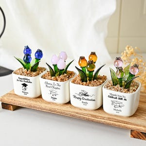 May include: Four square white ceramic pots containing glass flowers in blue, light pink, amber, and pink, each filled with small pebbles. The pots feature black printed graduation messages and names, arranged on a rectangular wooden base.