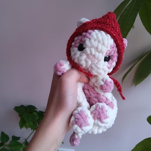 Puede incluir: Un gato de peluche tejido a crochet que lleva un gorro rojo tejido a crochet con un acabado brillante. El gato es blanco y rosa con ojos negros.