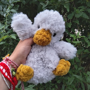 Puede incluir: Un peluche de pato gris con un pico y patas amarillas texturizadas. El juguete de peluche tiene pequeños ojos negros y alas esponjosas. El pato se sostiene frente a un fondo de hojas verdes.