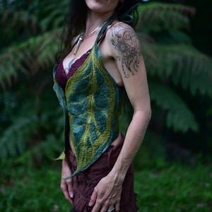 May include: A woman wearing a green and brown leaf-shaped top with a floral tattoo on her shoulder. She is standing in a forest setting.