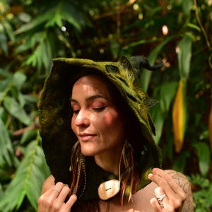 May include: A green hooded cloak with leaf and feather accents. The cloak features a large hood with a pointed tip and a wooden button closure. The woman in the image is wearing the cloak in a lush, green environment.