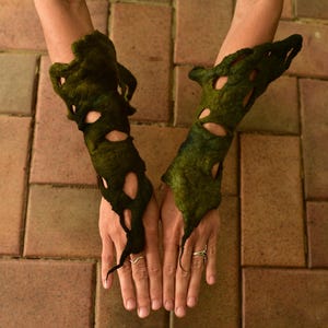 May include: Dark green felted arm cuffs with irregular cutouts. The cuffs are worn on the arms, with the hands visible. The cuffs have a textured appearance and are a unique accessory.
