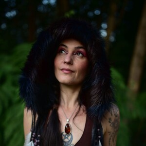 May include: A woman with long dark hair wearing a black fur-lined hooded jacket, a brown tank top, and a silver necklace with a large amber pendant. She has a tattoo on her left shoulder and is wearing a beaded and feather hair accessory.