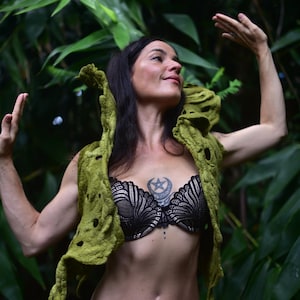 May include: A woman wearing a green felt vest with a unique, open design. The vest has a textured appearance and is paired with a black lace bra and purple leggings. The background features lush green foliage.