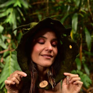 May include: A dark green felted hood with leaf-like cutouts. The hood is worn by a person with a wooden pendant necklace and feather accents. The background features lush green foliage, creating a natural, earthy aesthetic.