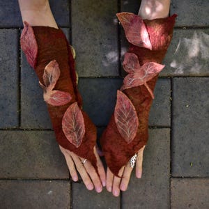 Op de afbeelding: Een paar bruine wollen armwarmers met bladontwerpen. De armwarmers zijn gemaakt van een natuurlijk materiaal en hebben een rustieke uitstraling.