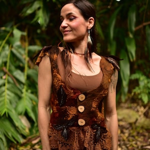 May include: A sleeveless waistcoat made of textured felted wool in shades of brown, rust, and orange. The garment features three large, round, light-coloured wooden buttons down the front, dark leaf-shaped appliqués, and a brown lace hem at the bottom.