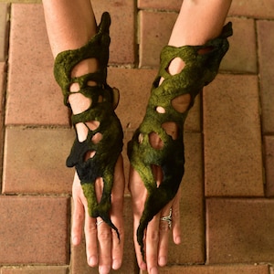 May include: A pair of unique, handmade arm cuffs. The cuffs are made of felted wool in shades of green and black, with an open, organic design. The cuffs are shown against a brick background.
