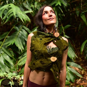 Puede incluir: Una mujer con un chaleco de fieltro verde con grandes botones de madera. El chaleco tiene un diseño único y texturizado con detalles recortados. Lleva leggings burdeos y tiene el pelo oscuro. El fondo es un exuberante follaje verde.