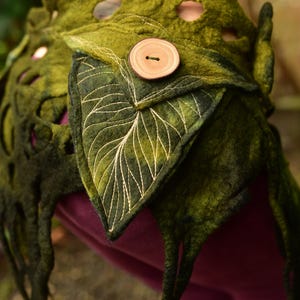 May include: A handcrafted, green felt leaf-shaped garment with a wooden button closure. The garment features intricate leaf vein embroidery and cut-out details. The design evokes a natural, organic aesthetic, suitable for costume or artistic wear.