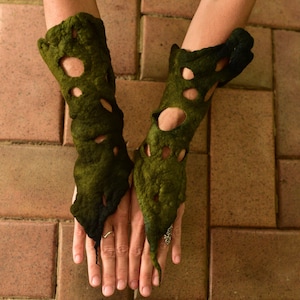 May include: A pair of olive green arm warmers with irregular cut-out holes. The textured, felted material covers the forearms and extends over the hands. The arm warmers are displayed on a brick surface.