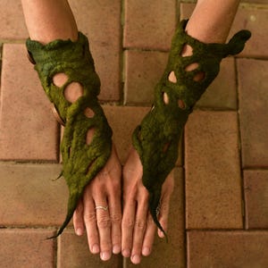 May include: Green felt arm cuffs with organic, leaf-like shapes and cut-out details. The cuffs are worn on the arms, with the hands visible. The cuffs have a textured appearance and are made of a dark green material.
