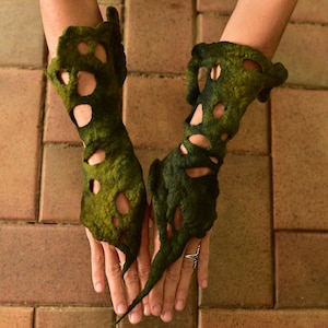 May include: A pair of unique, green felt gloves with irregular cut-out patterns. The gloves have a textured appearance and a dark green hue, with lighter green and tan accents. The gloves are displayed against a brick background.