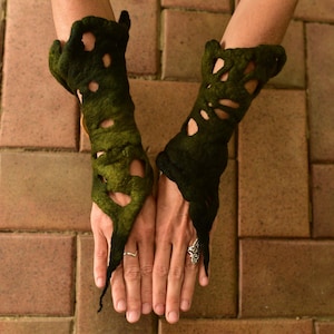May include: A pair of dark green felted arm cuffs with cut-out designs. The cuffs are worn on the wrists and extend down the hand. The cuffs are a dark green color and have a textured appearance.