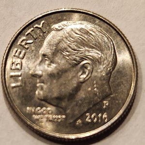 May include: A close-up of a United States dime coin. The coin features a profile of a man's head, the word "LIBERTY" at the top, and the year "2016" at the bottom. The coin is silver in color and has a textured surface.