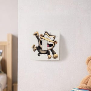 May include: A white light switch cover with a cartoon monkey design. The monkey, in shades of brown and tan, has a wide grin and is raising an arm. The light switch is in its mouth. The background is a white wall.