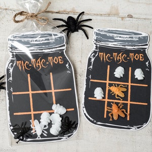 Puede incluir: Dos tableros de juego de tic-tac-toe de tarro de albañil en blanco y negro con las palabras "Tic-Tac-Toe" impresas en ellos. Los tableros están decorados con líneas naranjas y llenos de piezas de juego de calavera blanca y mosca naranja.