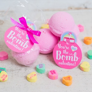 Puede incluir: Bombas de baño rosas con una etiqueta rosa y blanca que dice "You're the Bomb Valentine!" Las bombas de baño están rodeadas de pequeños caramelos en forma de corazón.