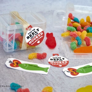 Puede incluir: Un recipiente de plástico transparente lleno de gusanos gomosos de colores y una etiqueta de San Valentín roja y blanca que dice "You've Reely Got Me Hooked Valentine."