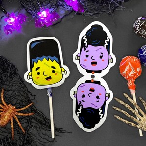 May include: Two Halloween-themed lollipop sticks with cartoon images of Frankenstein's monster. The top stick features a purple monster with black hair and the bottom stick features a yellow monster with blue hair. The sticks are on a gray surface with a spider, a skeleton hand, and candy.