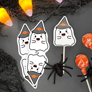 May include: Four white ghost-shaped Halloween decorations with black witch hats and smiling faces. The ghosts are on sticks and are surrounded by black netting, orange and white lights, and two orange lollipops with black spiders.