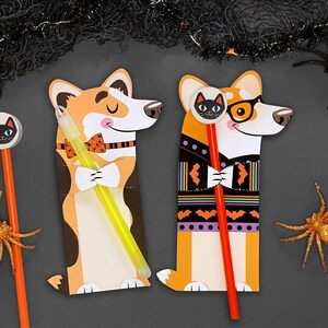 May include: Two Halloween-themed pencil holders shaped like corgis. One corgi is wearing a bow tie and holding a glow stick. The other corgi is wearing a sweater with bats and holding a red pencil.
