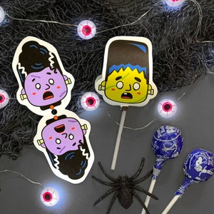 May include: Two Frankenstein monster face cutouts on sticks, one purple and one yellow, with a black spider and blue lollipops on a black and white background with pink and white string lights.