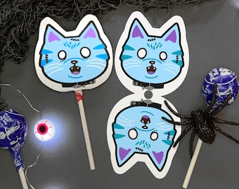 May include: Three blue cat face Halloween masks on sticks. The masks have purple ears, white eyes, and black mouths. The masks are on a gray background with candy and other Halloween decorations.