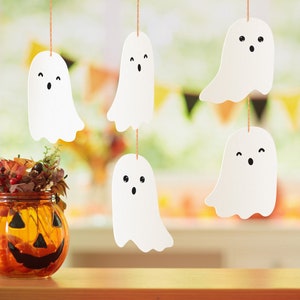 Könnte beinhalten: Fünf weiße Papiergeister mit schwarzen Augen und Mündern hängen von der Decke. Eine kleine orangefarbene Kürbislaterne steht auf einem Holztisch mit einer Vase mit Herbstblättern davor.