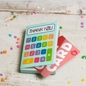 Peut inclure: Une carte en forme de calculatrice bleue et verte avec les mots "Thank You" sur l'écran. La carte est ouverte pour révéler une carte-cadeau rose avec le mot "CARD" imprimé dessus.