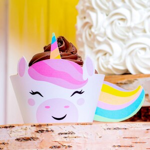 Op de afbeelding: Twee chocolade cupcakes versierd met cupcake-wikkelingen in de vorm van eenhoorns. De wikkelingen zijn wit met roze en gele eenhoornhoorns en een glimlachend eenhoorn gezicht. De cupcakes staan op een houten taartstandaard.