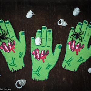 May include: Three green zombie hands with red blood and black spiders on a dark brown background.  The hands are decorated with white plastic skulls.