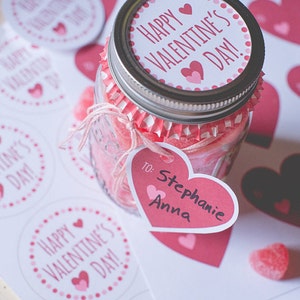 Op de afbeelding: Een glazen pot gevuld met roze hartvormige snoepjes, versierd met een roze en wit gepolst hartvormig etiket met de tekst "Happy Valentine's Day!" en een roze hartvormig label met de tekst "To: Stephanie Anna".