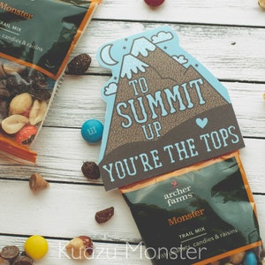 Könnte beinhalten: Eine blaue und braune Karte mit dem Text "To Summit Up You're The Tops" und einer Bergillustration. Die Karte ist an einer Tüte Trailmix mit dem Markennamen "Archer Farms" und dem Produktnamen "Monster Trail Mix" befestigt.