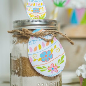 May include: A clear glass mason jar with a silver lid and a brown twine bow. The jar is filled with layers of brown, white, and tan ingredients. A colourful Easter egg-shaped paper tag with a floral design is tied to the jar with the twine. The jar is labelled "MASON".