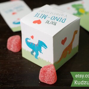 Puede incluir: Una peque&ntilde;a caja de regalo blanca con un coraz&oacute;n rojo y el texto "You're Dino-Mite!" La caja tiene una ilustraci&oacute;n de dinosaurio en la parte delantera y est&aacute; decorada con corazones rojos.
