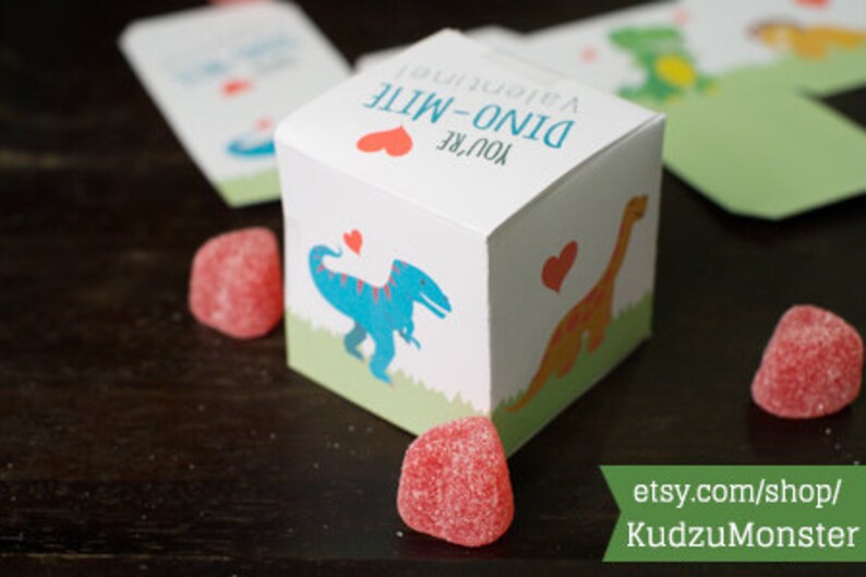 Puede incluir: Una peque&ntilde;a caja de regalo blanca con un coraz&oacute;n rojo y el texto "You're Dino-Mite!" La caja tiene una ilustraci&oacute;n de dinosaurio en la parte delantera y est&aacute; decorada con corazones rojos.
